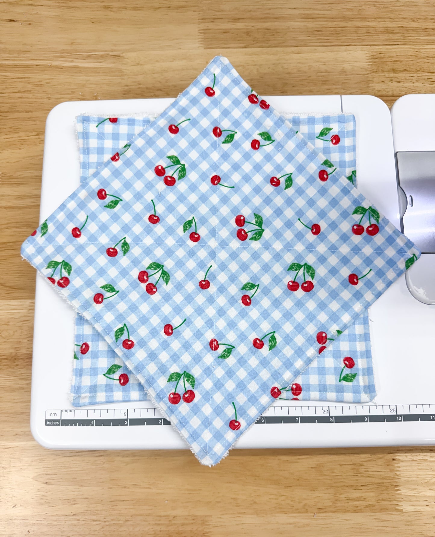 Two square fabric items, likely napkins or small table mats, stacked on a white sewing machine. The top fabric has a light blue and white gingham check pattern with a repeating print of red cherries with green leaves.