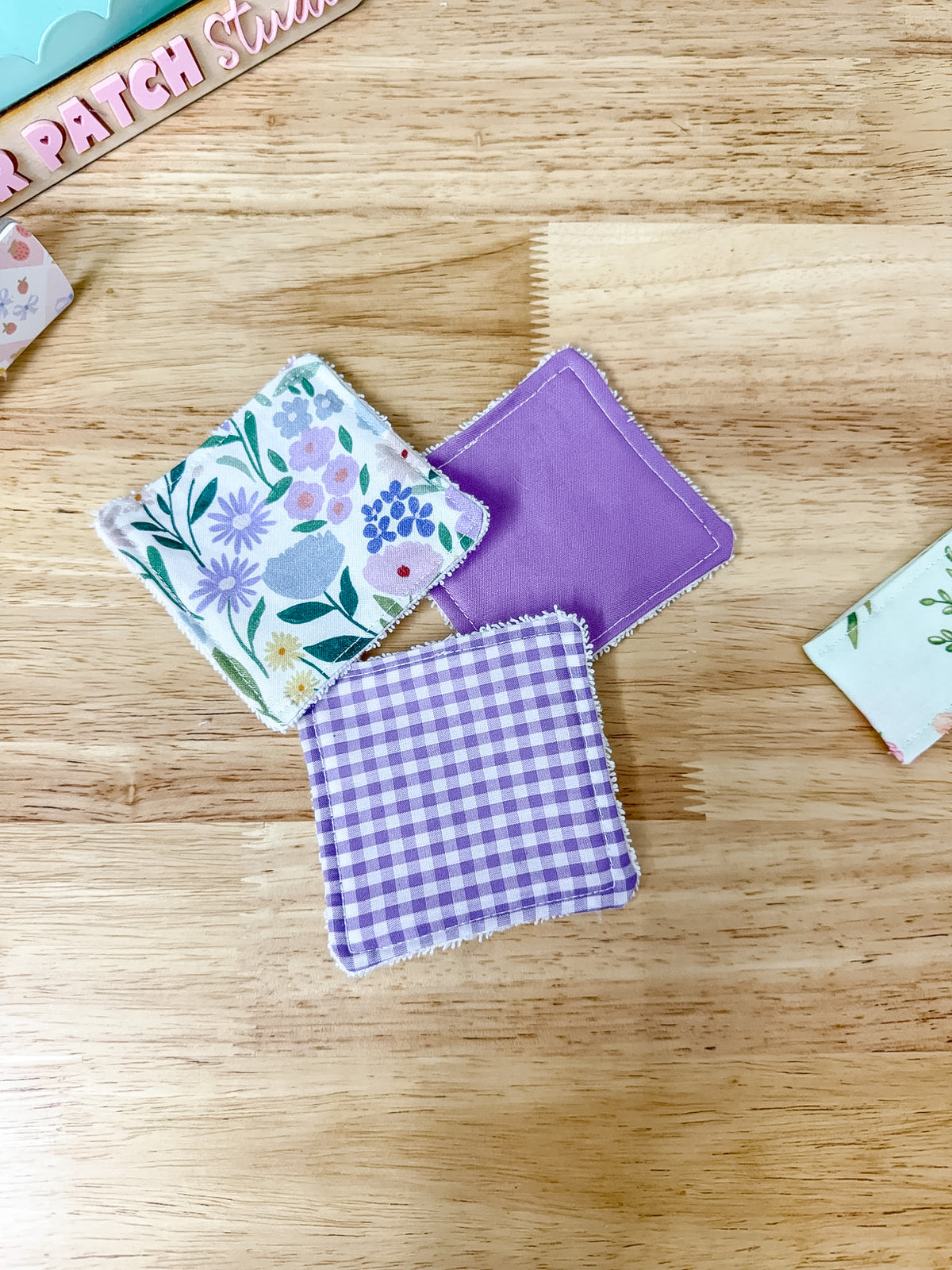 Three small fabric square patches with frayed edges are arranged on a light wood surface. One patch has a floral print in pastel colors, one is solid lavender purple, and one is a purple-and-white gingham pattern.