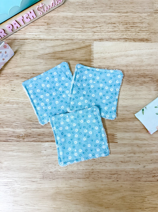 Three small square fabric coasters with a light blue background and white floral pattern, arranged on a light wood tabletop.