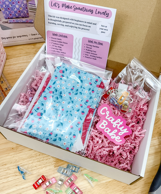 An open gift box on a wooden surface containing a craft kit with a light blue floral fabric pouch, pink shredded filler, a pink 'Crafty Babe' tag, and an instruction card reading 'Let's Make Something Lovely.'.