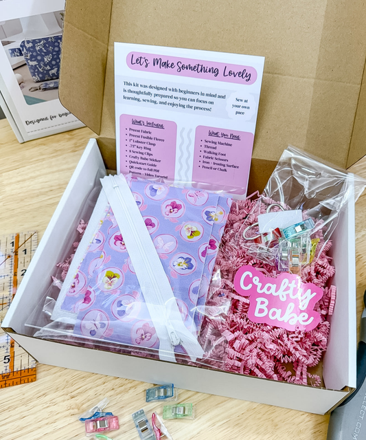 An open white box filled with craft supplies and a printed card reading 'Let's Make Something Lovely.' Visible items include a lavender patterned bag, pink shredded paper filler, a pink 'Crafty Babe' tag, small clear and pastel clips, and a kraft cardboard insert.