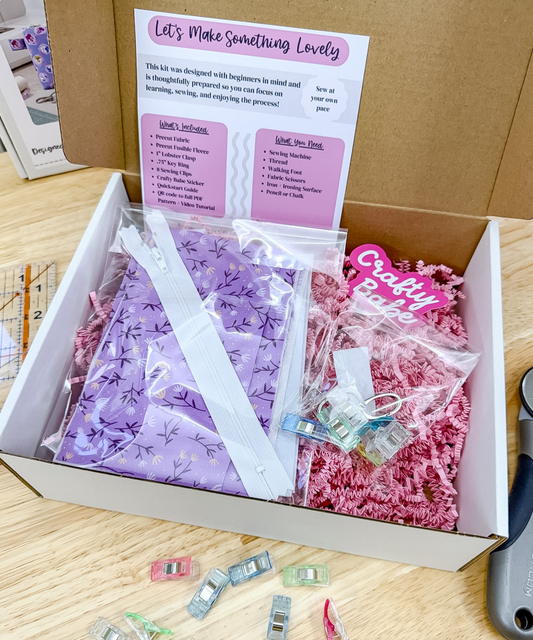 An open cardboard craft kit box on a wooden surface, filled with purple patterned paper, pink shredded paper, clear plastic bags, small clips, and a printed instruction card. A hand is visible holding the box edge, so this is classified as Lifestyle rather than Product/Packaging.