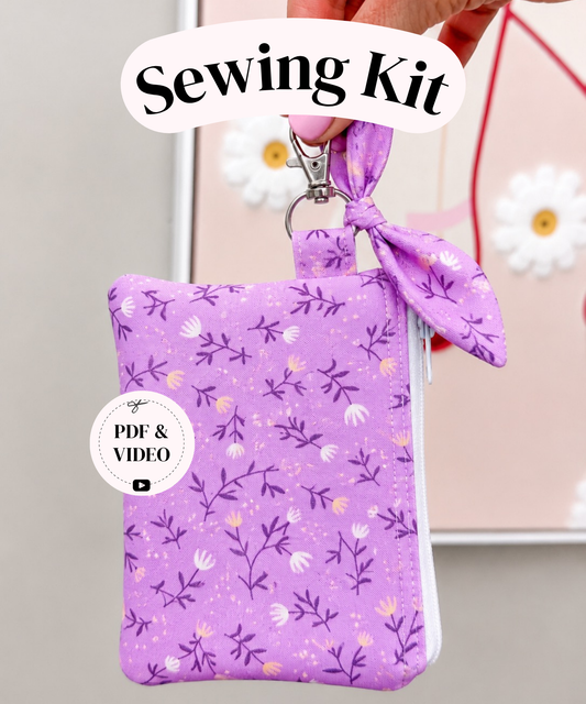 A purple fabric sewing kit pouch with a floral print, held by a hand. The pouch has a matching fabric wrist loop and a metal clasp.