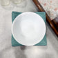 White bowl on a teal placemat with a floral tablecloth in the background