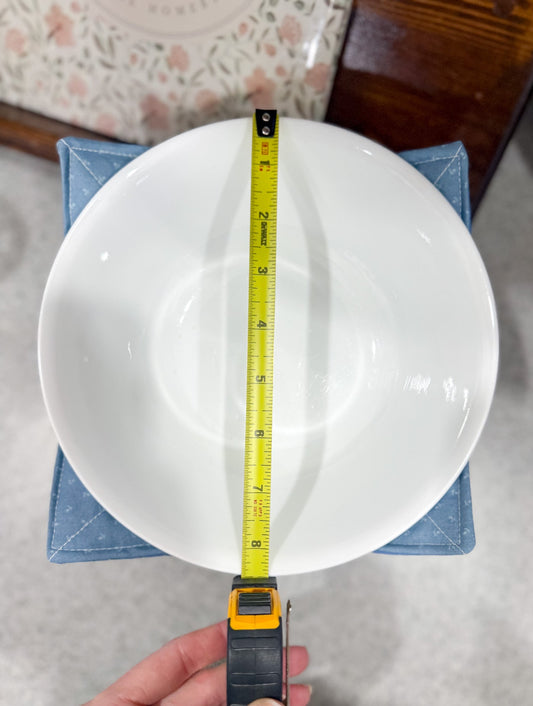 White bowl with a measuring tape on top, held by a hand, against a neutral background.
