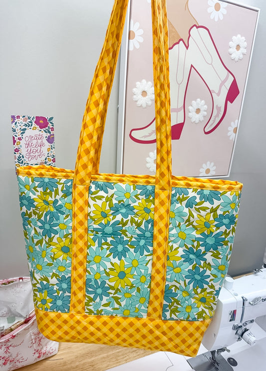 Yellow and floral-patterned tote bag on a sewing machine with a sewing-themed wall art in the background.