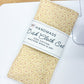 A folded dish cloth set with a yellow and orange floral pattern, wrapped in a white paper band that reads 'HANDMADE Dish Cloth Set 100% COTTON MACHINE WASH & DRY,' resting on a white cutting mat over a light wood table.