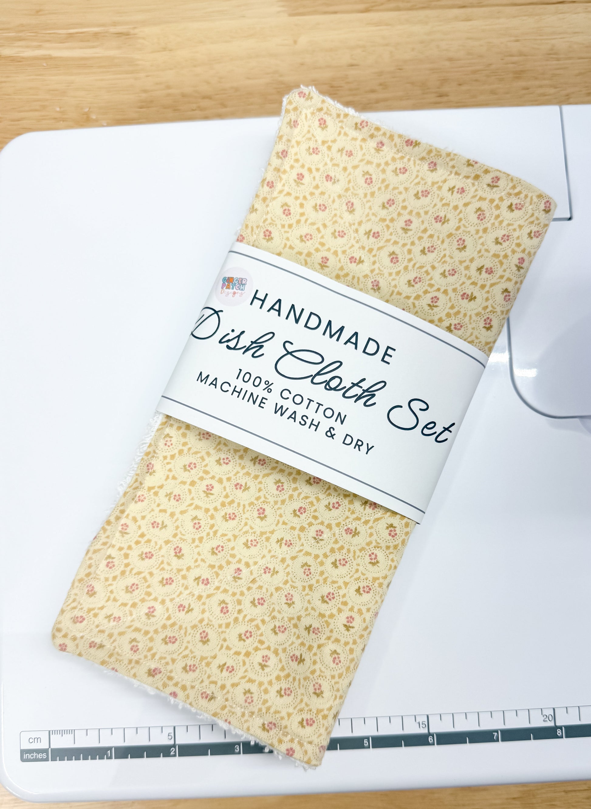 A folded dish cloth set with a yellow and orange floral pattern, wrapped in a white paper band that reads 'HANDMADE Dish Cloth Set 100% COTTON MACHINE WASH & DRY,' resting on a white cutting mat over a light wood table.