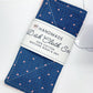 A folded blue dishcloth set with a small pink and white pattern, wrapped in a white paper band that reads 'HANDMADE Dish Cloth Set 100% COTTON MACHINE WASH & DRY,' placed on a white sewing machine over a light wood surface.