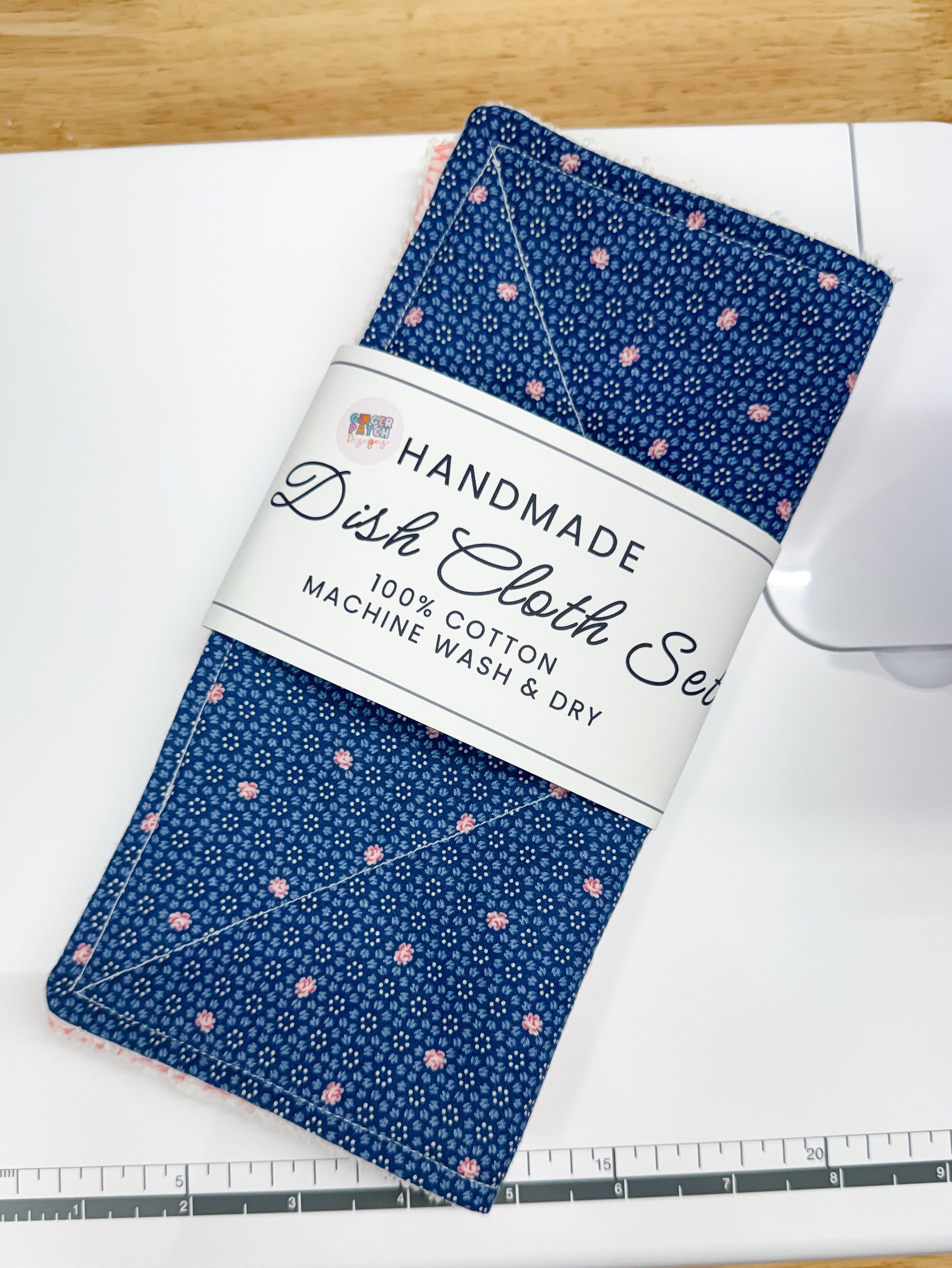 A folded blue dishcloth set with a small pink and white pattern, wrapped in a white paper band that reads 'HANDMADE Dish Cloth Set 100% COTTON MACHINE WASH & DRY,' placed on a white sewing machine over a light wood surface.