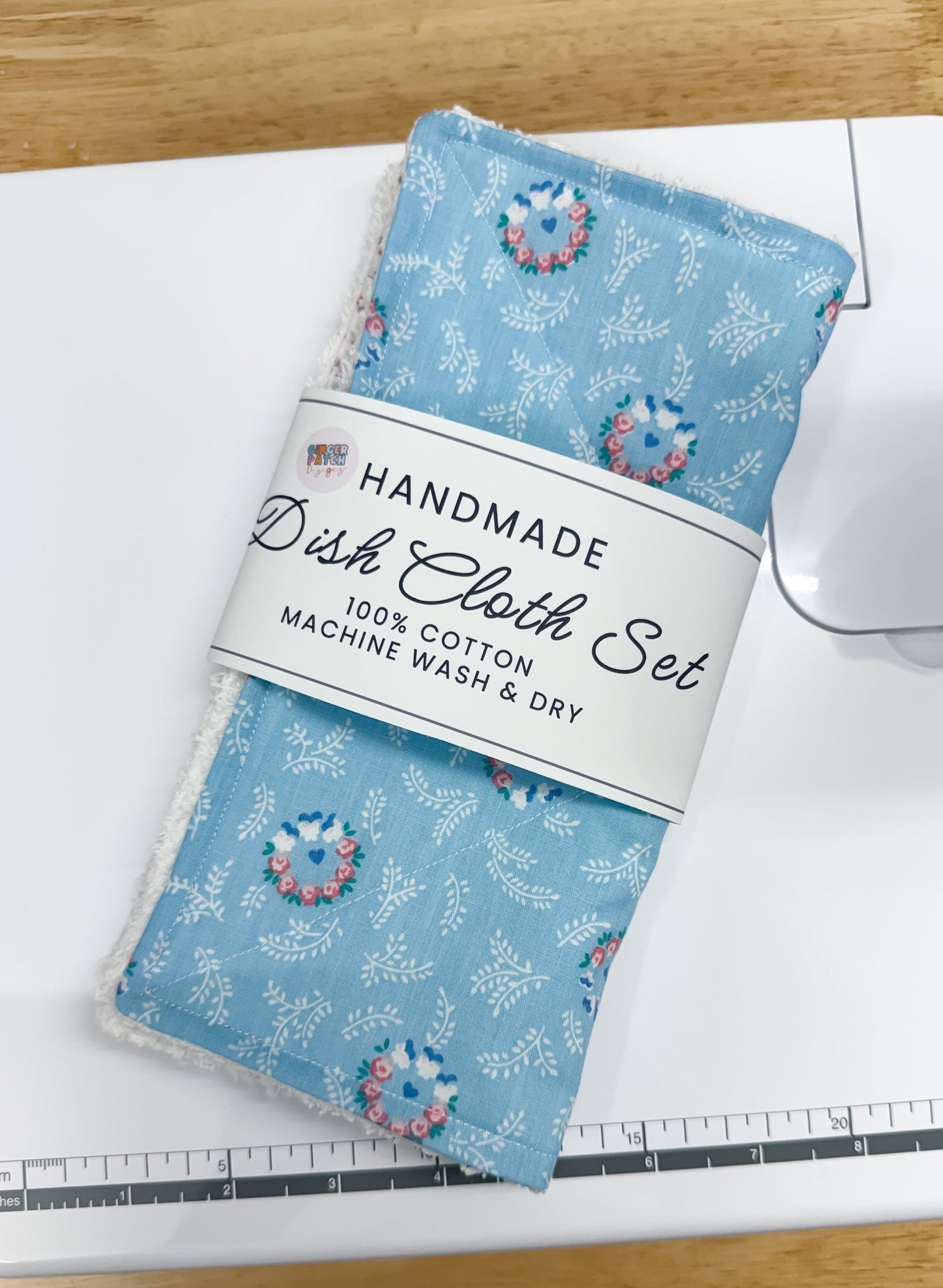 A handmade dish cloth set in a light blue floral print, wrapped in a white paper band label and placed on top of a white sewing machine over a wooden table.