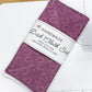 A handmade dish cloth set in purple with a white patterned band and a white paper label, placed on a white sewing machine over a light wood table.