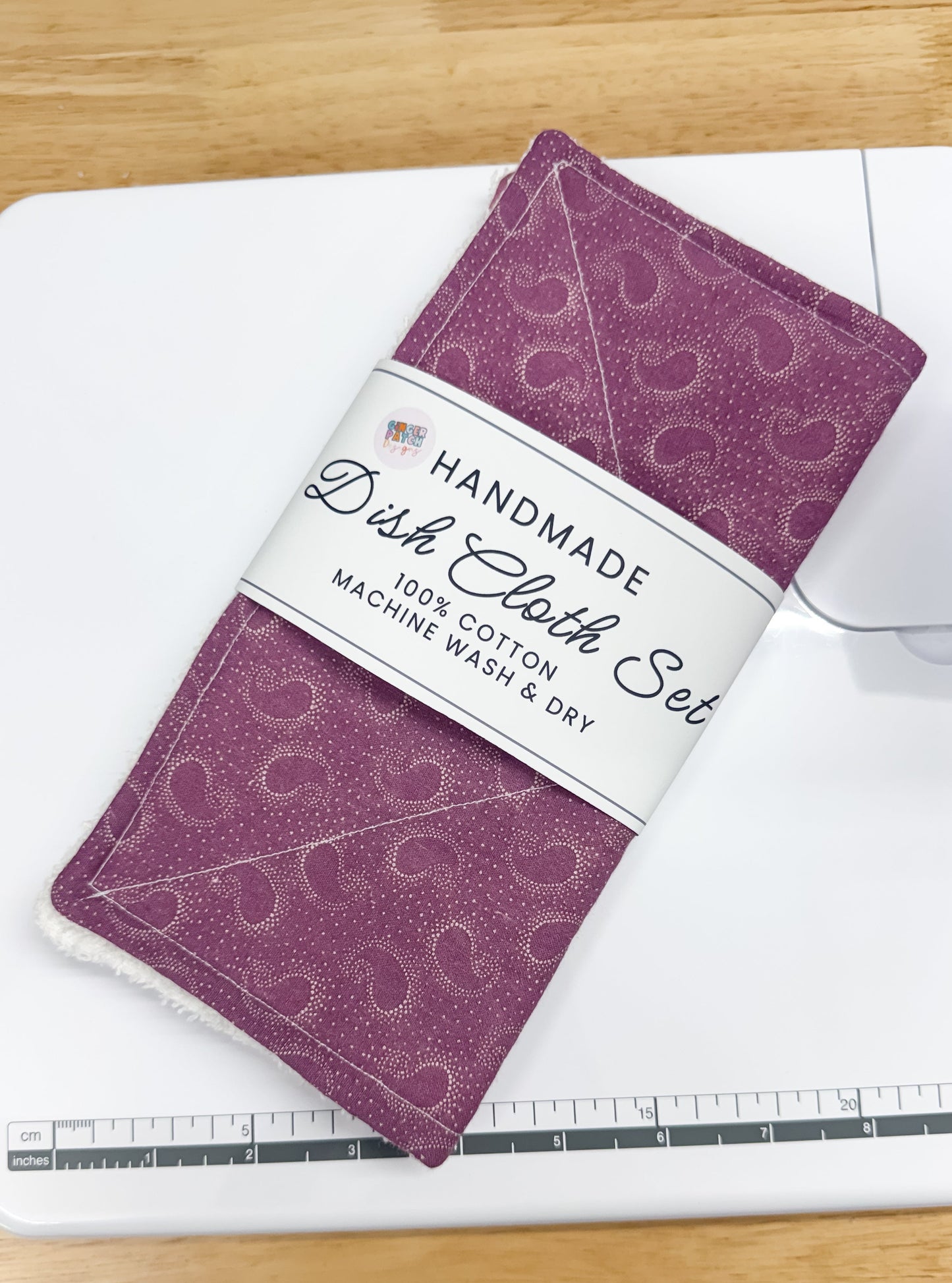 A handmade dish cloth set in purple with a white patterned band and a white paper label, placed on a white sewing machine over a light wood table.