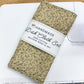 A handmade dish cloth set with a beige and green speckled pattern, wrapped in a white paper band labeled 'HANDMADE Dish Cloth Set 100% COTTON MACHINE WASH & DRY', resting on a white sewing machine over a light wood table.