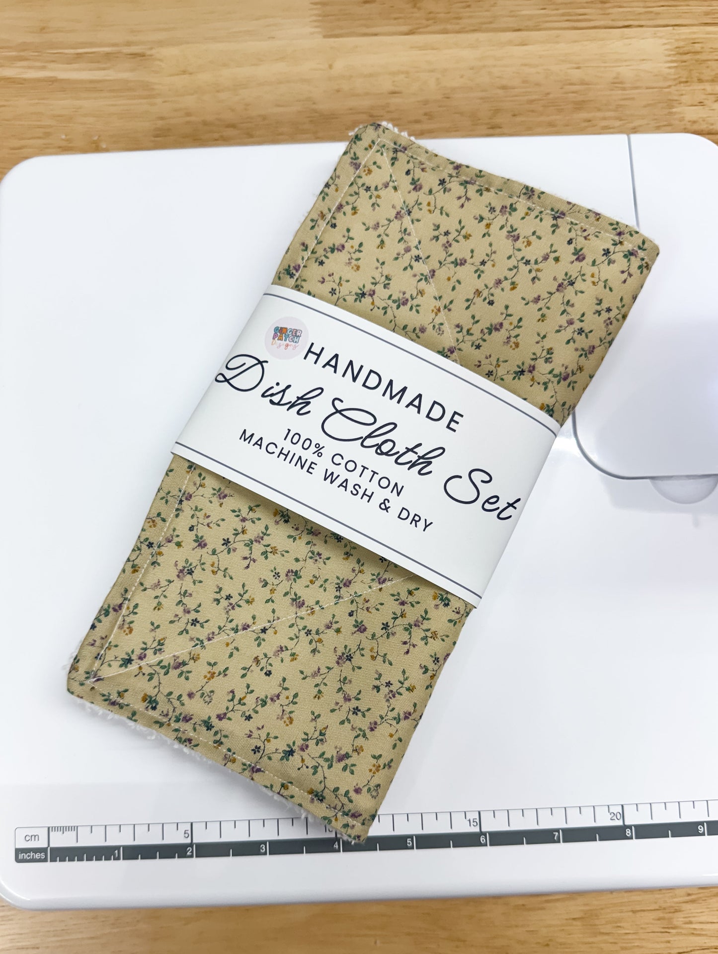 A handmade dish cloth set with a beige and green speckled pattern, wrapped in a white paper band labeled 'HANDMADE Dish Cloth Set 100% COTTON MACHINE WASH & DRY', resting on a white sewing machine over a light wood table.