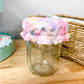 A clear glass mason jar with a floral fabric cover on top, placed on a light wood tabletop with a wicker basket and pastel decor in the background.