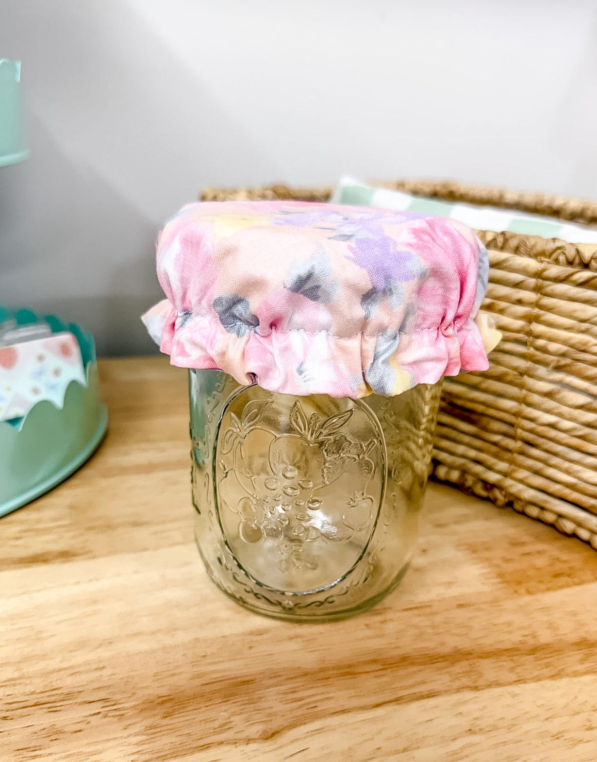 A clear glass mason jar with a floral fabric cover on top, placed on a light wood tabletop with a wicker basket and pastel decor in the background.