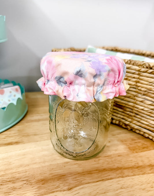 A clear glass mason jar with a floral fabric cover on top, placed on a light wood tabletop with a wicker basket and pastel decor in the background.