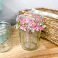 A clear glass mason jar with a pink and green floral patterned fabric lid, placed on a light wood tabletop next to another empty glass jar and a woven basket.