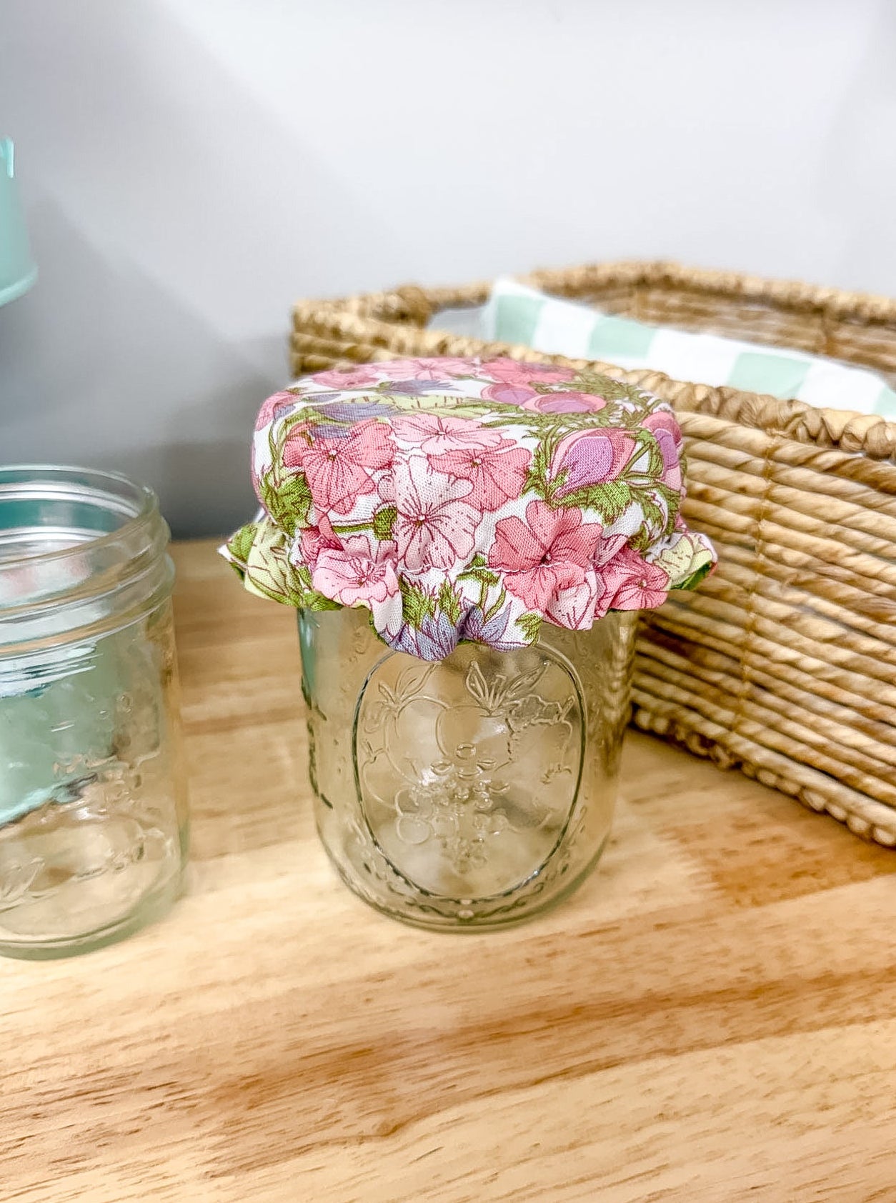 A clear glass mason jar with a pink and green floral patterned fabric lid, placed on a light wood tabletop next to another empty glass jar and a woven basket.