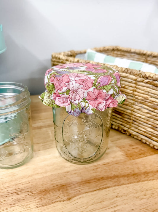 A clear glass mason jar with a pink and green floral patterned fabric lid, placed on a light wood tabletop next to another empty glass jar and a woven basket.