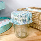 A clear glass mason jar with a ruffled fabric cover in a blue and white floral toile pattern, set on a light wood tabletop next to a woven basket.