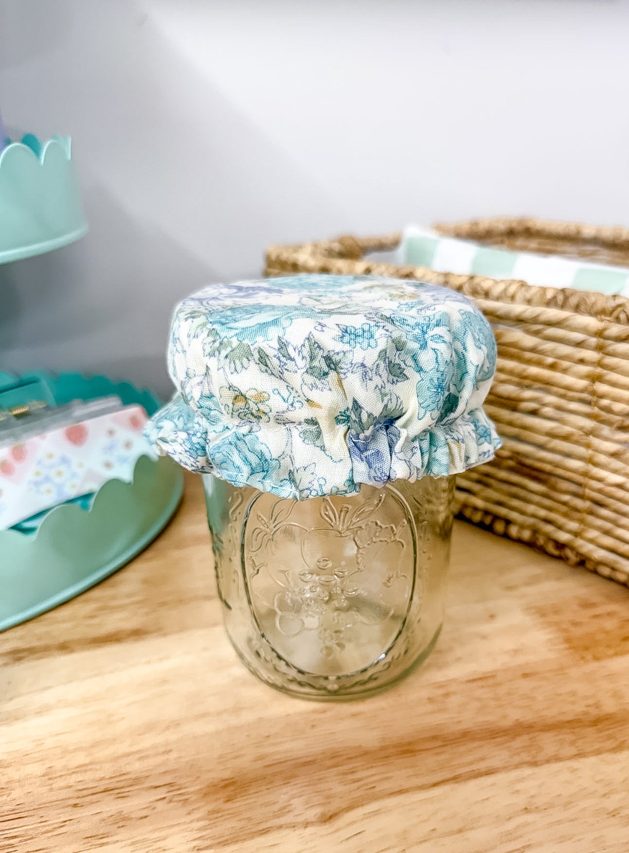 A clear glass mason jar with a ruffled fabric cover in a blue and white floral toile pattern, set on a light wood tabletop next to a woven basket.