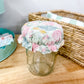 A clear glass mason jar with a floral fabric cover on top, placed on a wooden tabletop next to a woven basket.