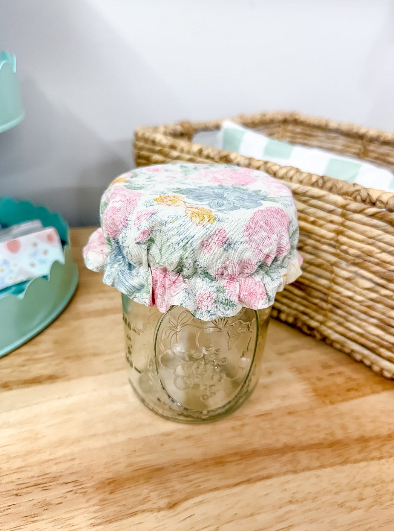 A clear glass mason jar with a floral fabric cover on top, placed on a wooden tabletop next to a woven basket.