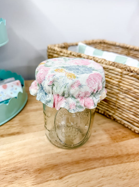 A clear glass mason jar with a floral fabric cover on top, placed on a wooden tabletop next to a woven basket.