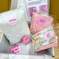 A gift box filled with baking and sewing supplies, including a large bag of white flour labeled 'Baking Q', pink patterned fabric pieces labeled 'Exterior Pieces' and 'Interior Pieces', a floral fabric piece labeled 'Batting Q', a printed booklet, and a pink 'Crafty Bake' sticker.