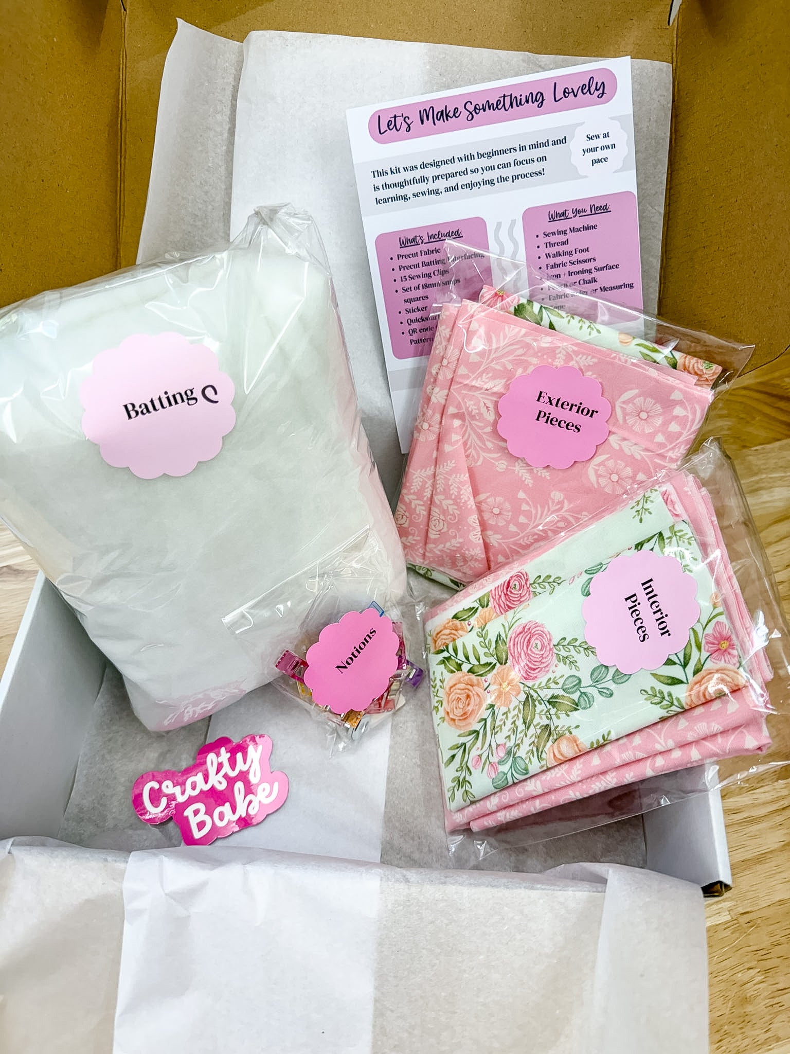 A gift box filled with baking and sewing supplies, including a large bag of white flour labeled 'Baking Q', pink patterned fabric pieces labeled 'Exterior Pieces' and 'Interior Pieces', a floral fabric piece labeled 'Batting Q', a printed booklet, and a pink 'Crafty Bake' sticker.