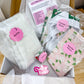 An open gift box filled with packaged baking or craft supplies, including a large white bag labeled 'Baking,' a pink floral packet labeled 'Exterior Pieces,' a pink floral packet labeled 'Emotional Pieces,' a small card reading 'Let's Make Something Lovely,' and a pink 'Crafty Bakes' sticker or label. The box is photographed on a wooden surface.