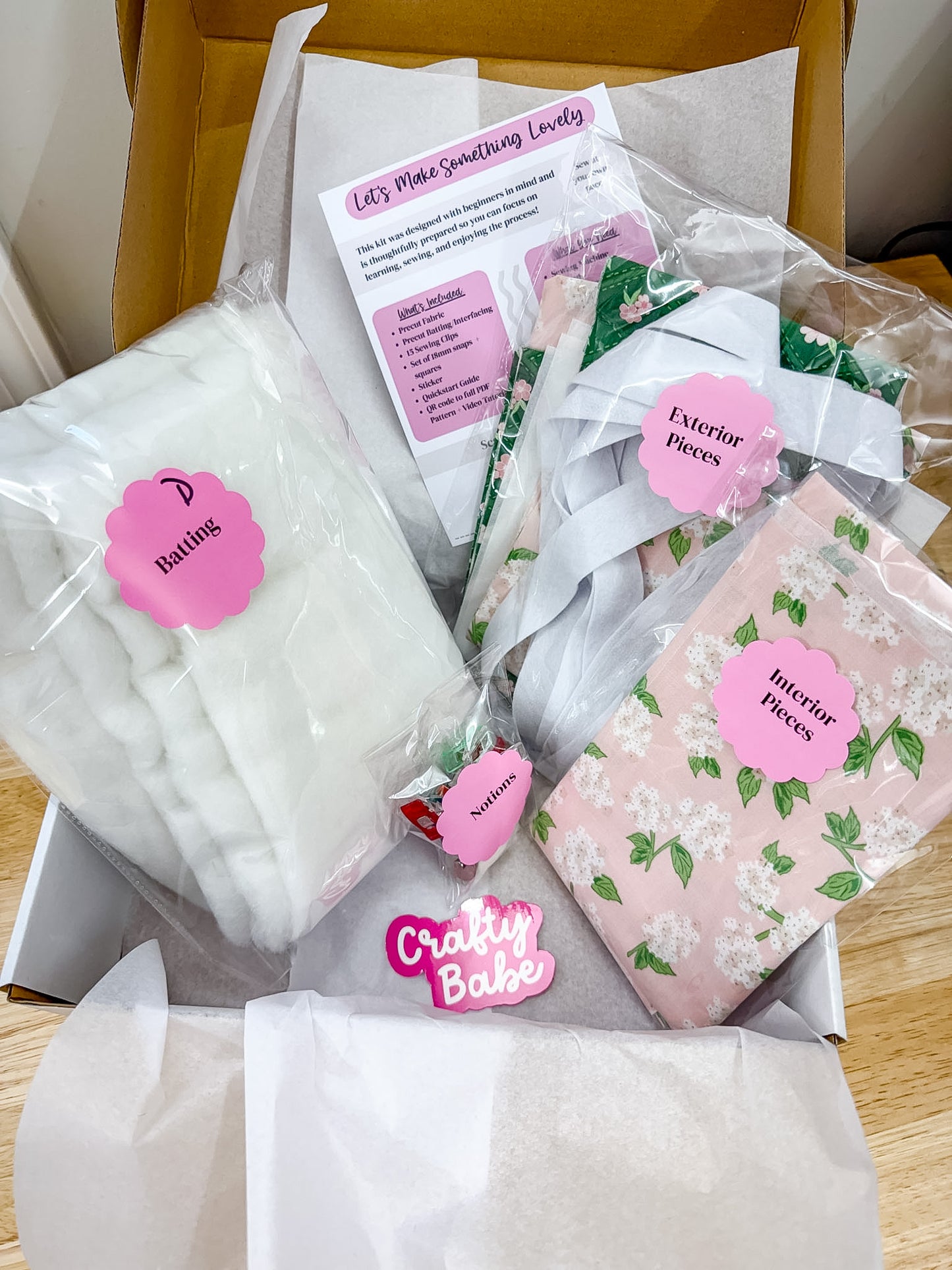 An open gift box filled with packaged baking or craft supplies, including a large white bag labeled 'Baking,' a pink floral packet labeled 'Exterior Pieces,' a pink floral packet labeled 'Emotional Pieces,' a small card reading 'Let's Make Something Lovely,' and a pink 'Crafty Bakes' sticker or label. The box is photographed on a wooden surface.