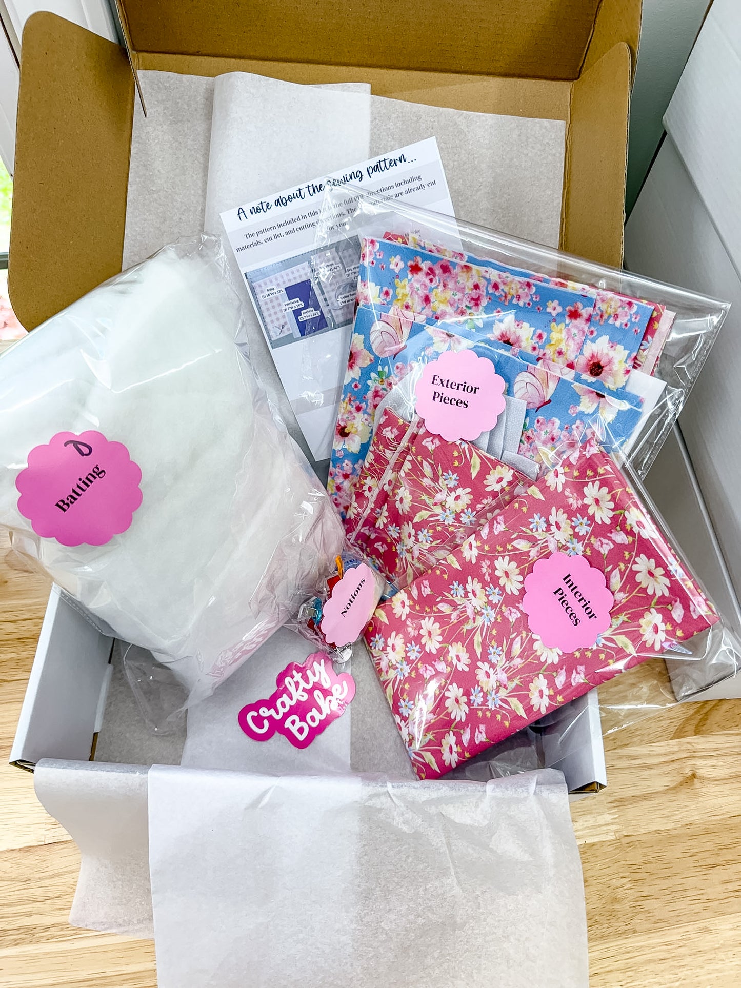A cardboard box filled with sewing supplies, including a large white bag labeled 'Bulbs', floral fabric pieces in blue and pink, and a pink tag reading 'Crafty Baker'.