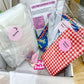 A baby gift box filled with baby items including a white bath towel, a red and white gingham blanket, a blue floral fabric piece, and a card that reads 'Let's Make Something Lovely'.