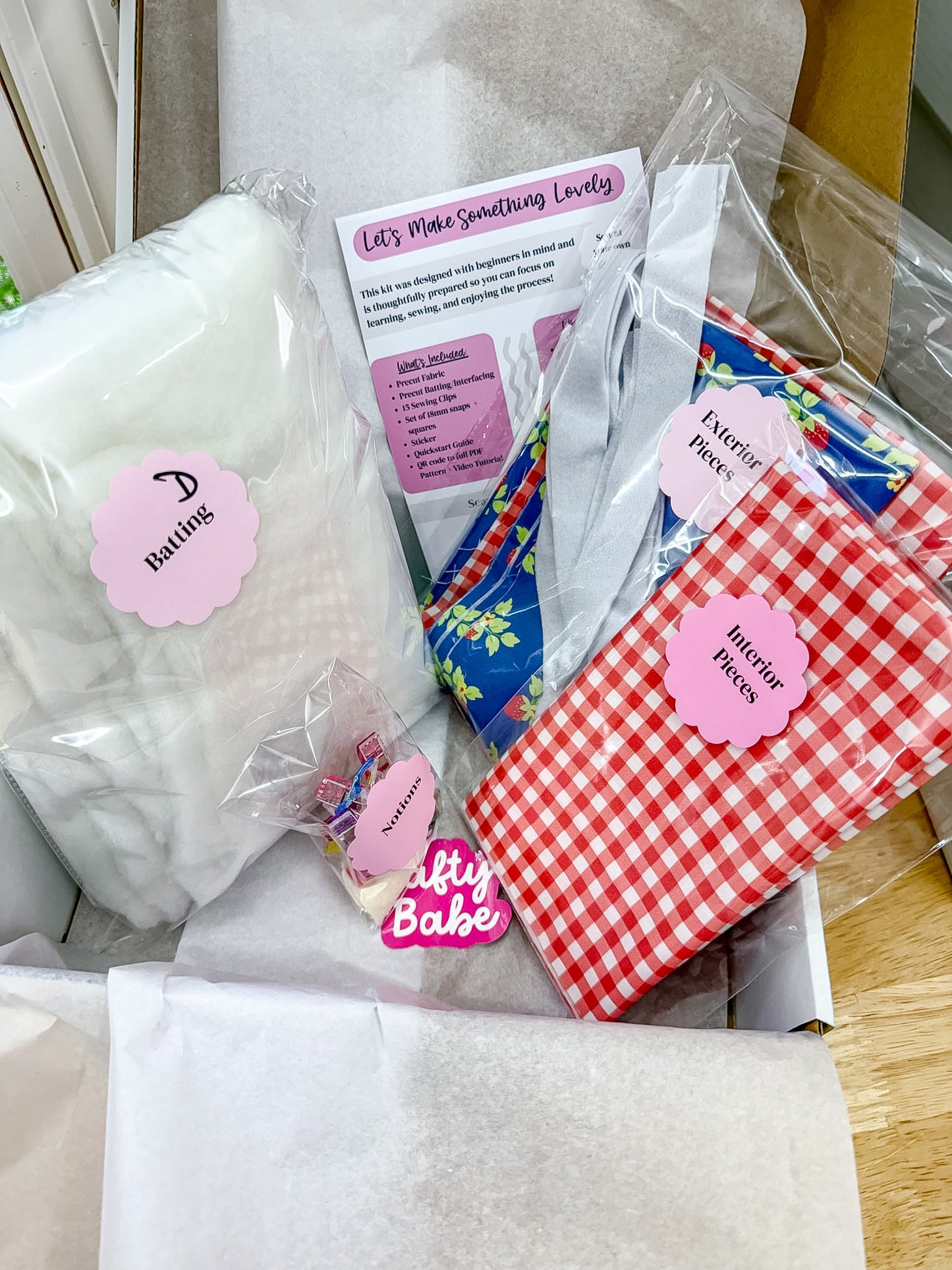 A baby gift box filled with baby items including a white bath towel, a red and white gingham blanket, a blue floral fabric piece, and a card that reads 'Let's Make Something Lovely'.