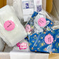 A sewing pattern kit in a cardboard box, including a white fabric piece labeled 'Basting,' blue floral fabric pieces labeled 'Exterior Backs' and 'Interior Backs,' a pink 'Crafty Babe' sticker, and a printed instruction sheet.