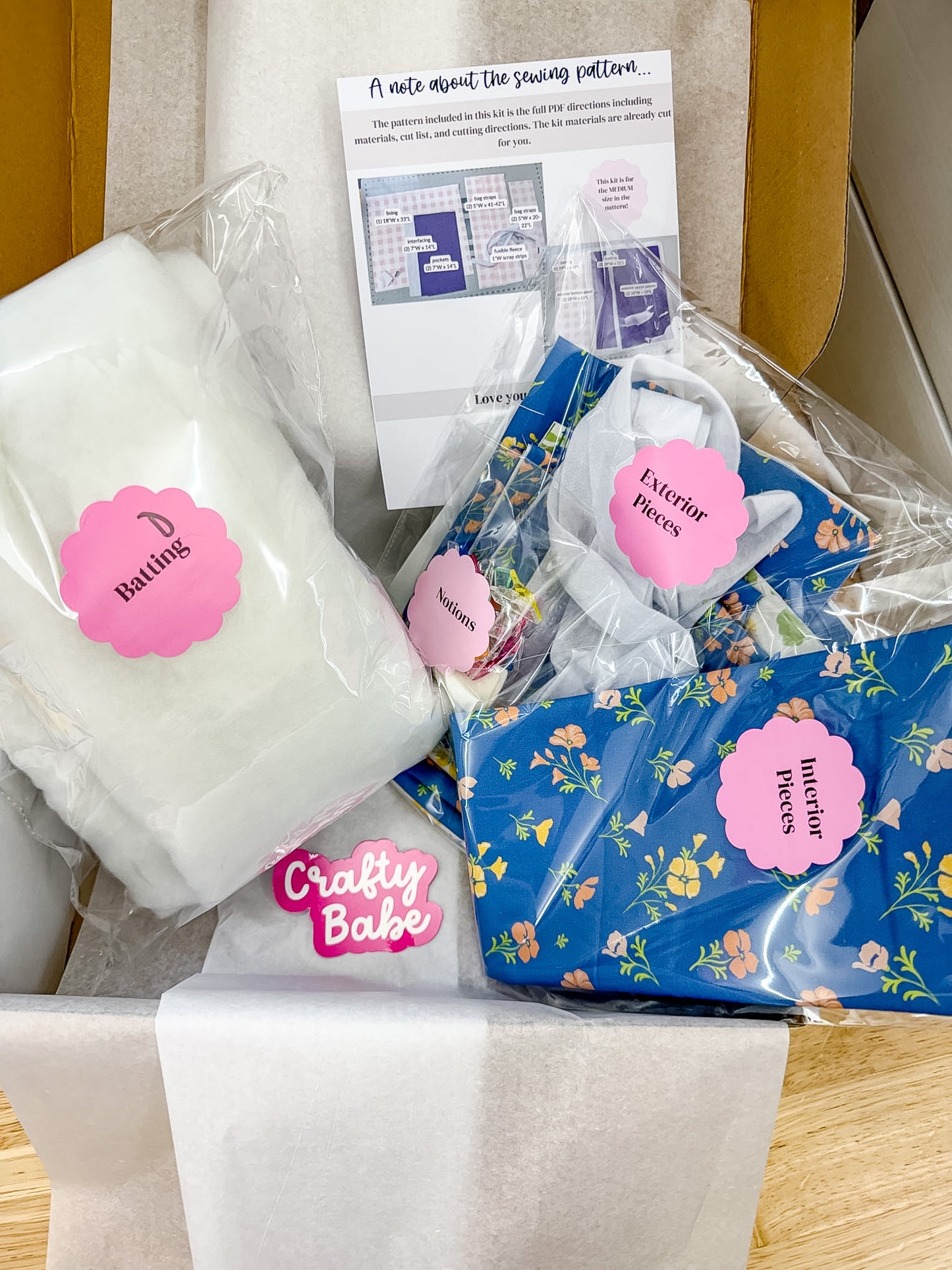 A sewing pattern kit in a cardboard box, including a white fabric piece labeled 'Basting,' blue floral fabric pieces labeled 'Exterior Backs' and 'Interior Backs,' a pink 'Crafty Babe' sticker, and a printed instruction sheet.