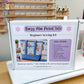 A boxed sewing kit labeled 'Daisy Mae Pocket Tote Beginner Sewing Kit' sits on a wooden table in front of a white sewing machine. The package shows fabric tote examples and a list of included items.