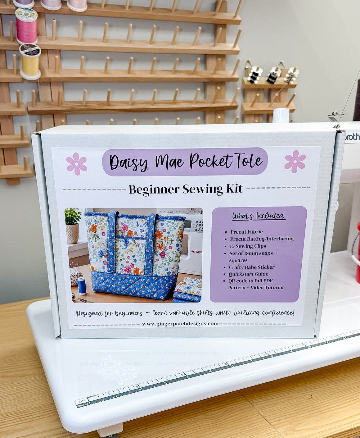 A boxed sewing kit labeled 'Daisy Mae Pocket Tote Beginner Sewing Kit' sits on a wooden table in front of a white sewing machine. The package shows fabric tote examples and a list of included items.