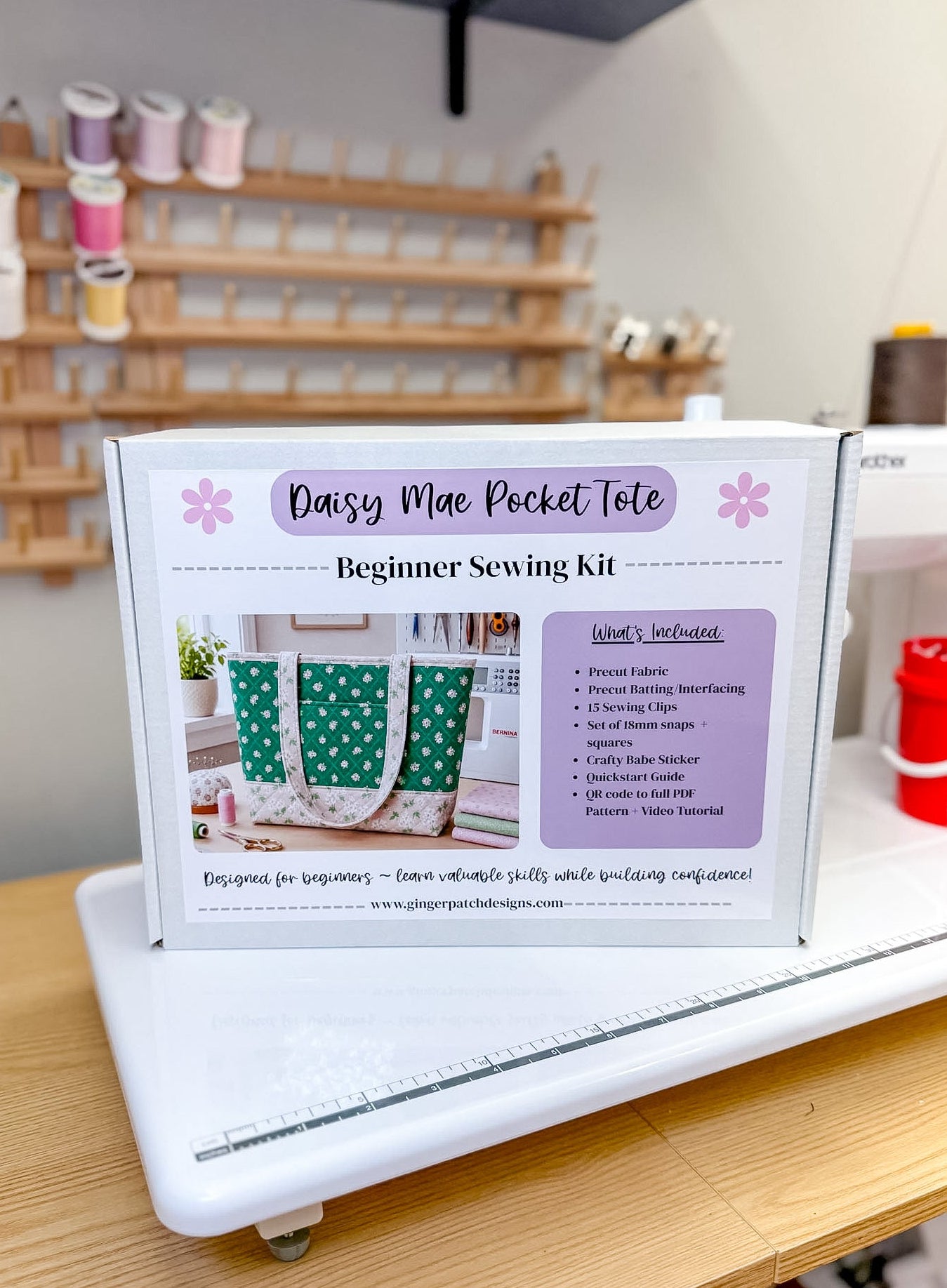 A boxed beginner sewing kit labeled 'Daisy Mae Pocket Tote' standing on a white machine on a wooden table, with a sewing supply rack blurred in the background.