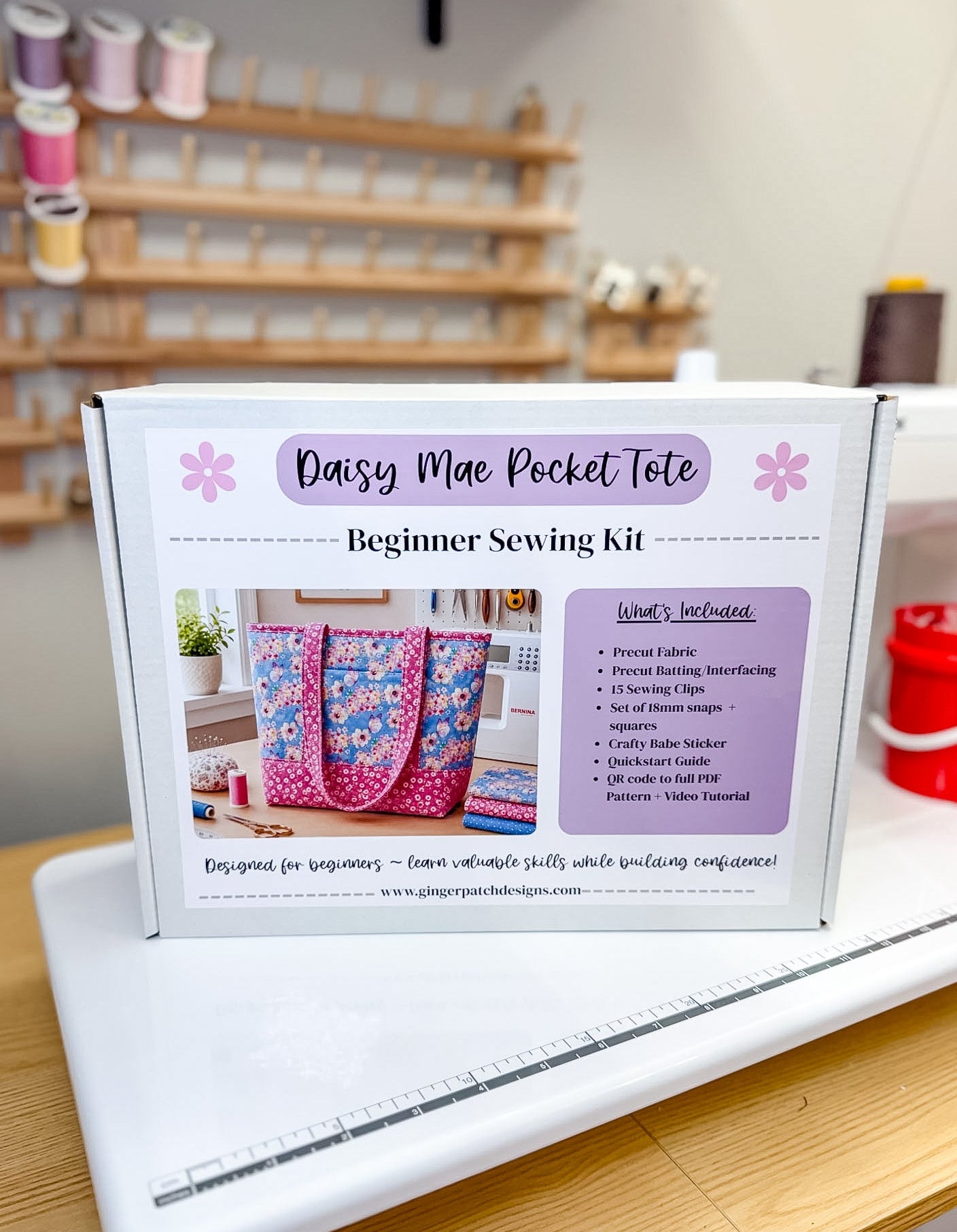 A white box labeled 'Daisy Mae Pocket Tote Beginner Sewing Kit' sits on a white sewing machine in a sewing room. The box shows a photo of a colorful quilted tote bag and lists included items such as pre-cut fabric, interfacing, sewing tips, and patterns.