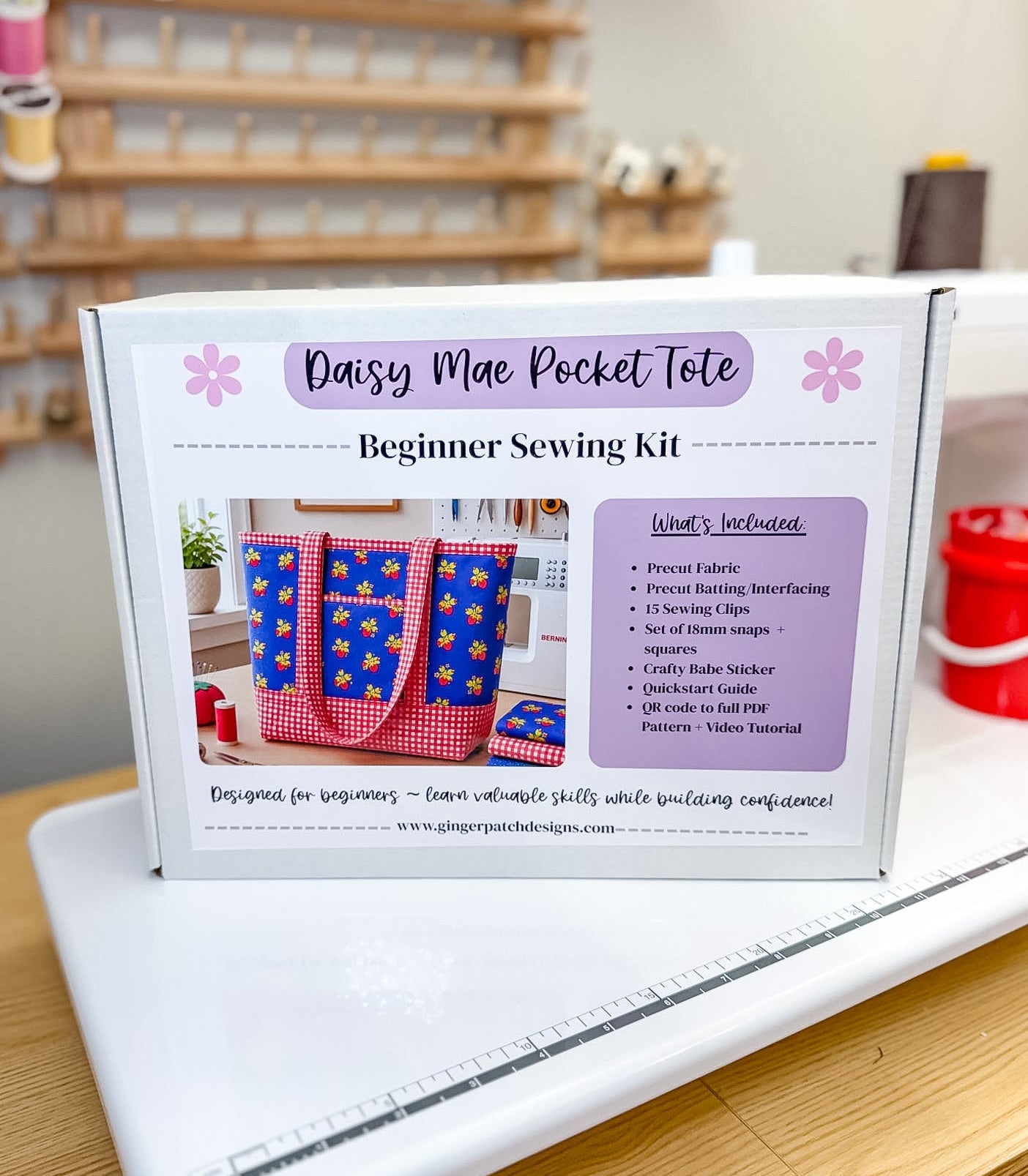 A white box labeled 'Daisy Mae Pocket Tote Beginner Sewing Kit' sits on a white sewing machine on a wooden table, with a blurred sewing room background containing shelves of thread.