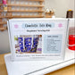 A white sewing kit box labeled 'Charlotte Tote Bag Beginner Sewing Kit' standing on a white sewing machine on a wooden table, with a sewing supply rack blurred in the background.