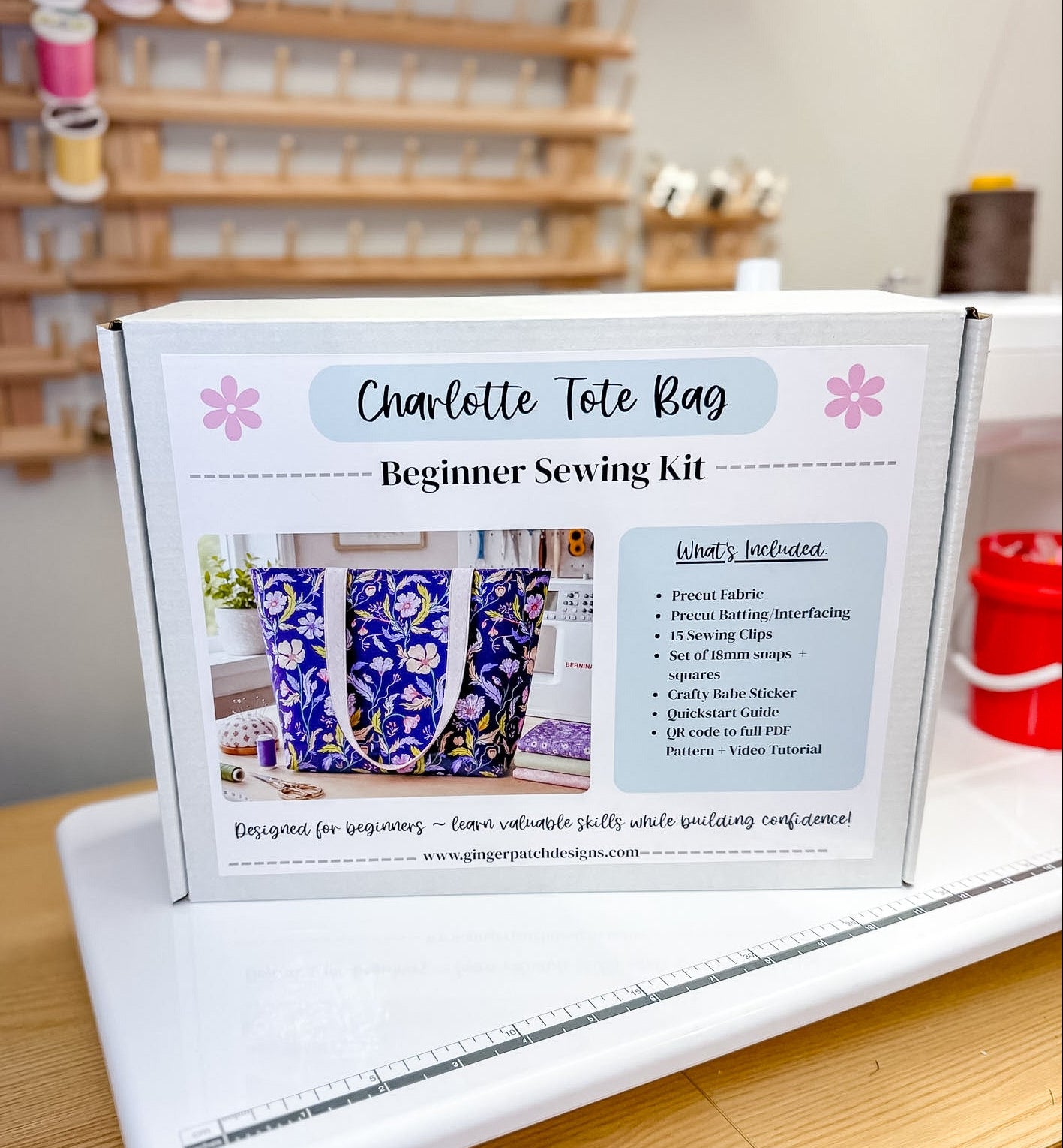 A white sewing kit box labeled 'Charlotte Tote Bag Beginner Sewing Kit' standing on a white sewing machine on a wooden table, with a sewing supply rack blurred in the background.