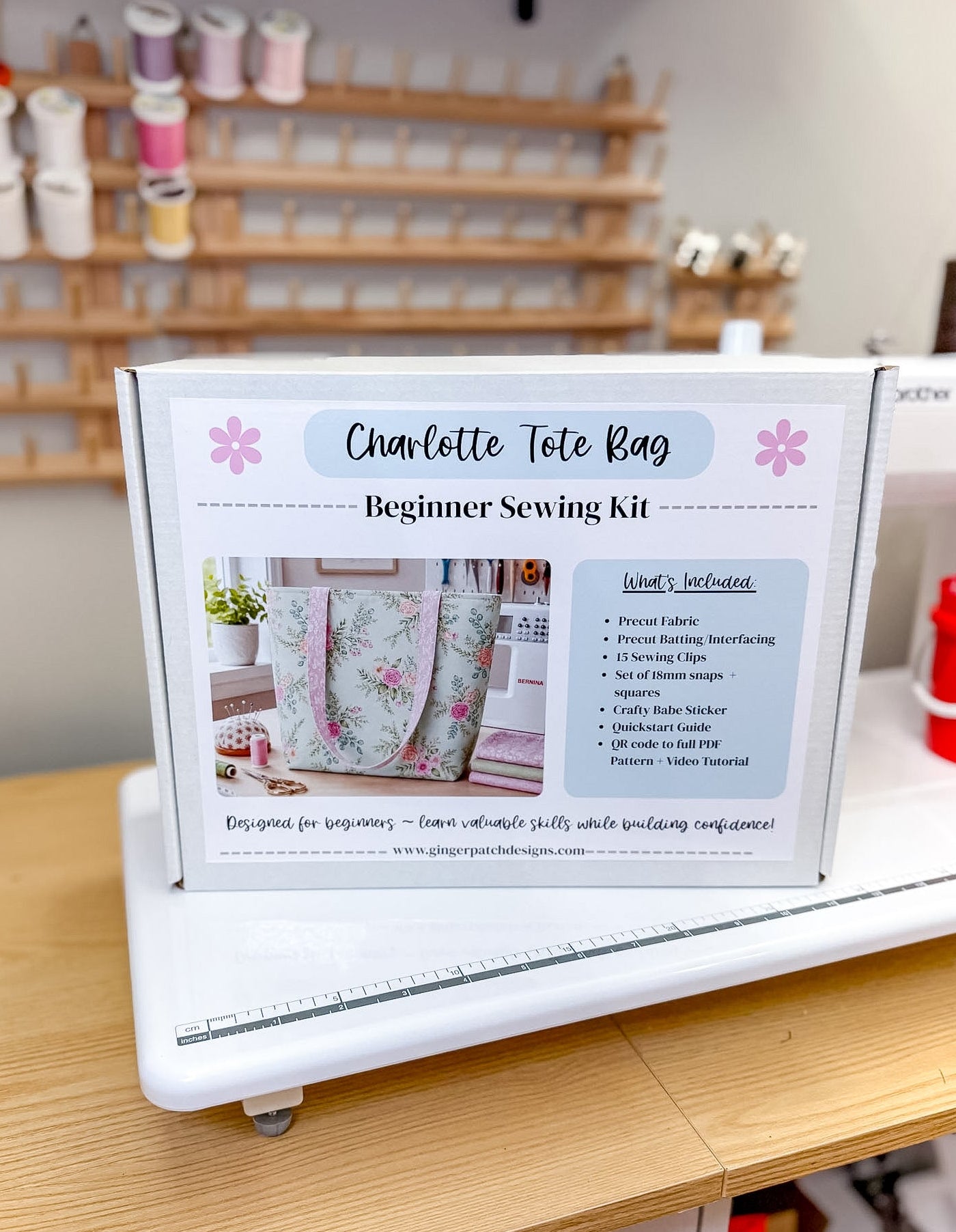A sewing kit box labeled 'Charlotte Tote Bag Beginner Sewing Kit' displayed on a white sewing machine in a bright craft room with shelves of thread in the background.