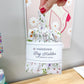 A hand with red nail polish holds a white hanging bag holder for grocery bags with a delicate multicolor floral print. The bag holder has a white paper band around the middle that reads 'HANGING Bag Holder FOR GROCERY BAGS'.
