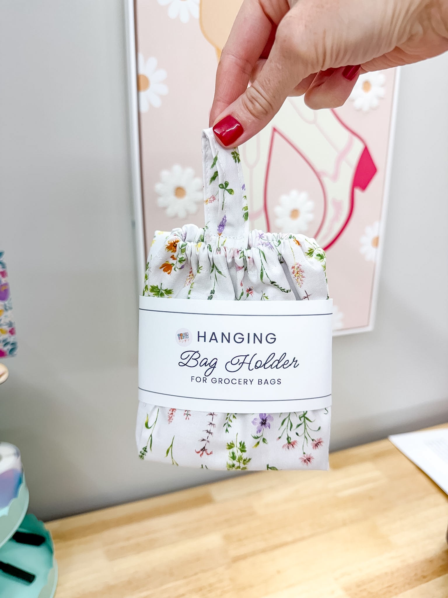 A hand with red nail polish holds a white hanging bag holder for grocery bags with a delicate multicolor floral print. The bag holder has a white paper band around the middle that reads 'HANGING Bag Holder FOR GROCERY BAGS'.