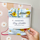A hand holds a light blue fabric hanging bag holder with a yellow sunflower print and a white paper label that reads 'Hanging Bag Holder for Grocery Bags.' The background shows a beige wall with framed floral artwork.